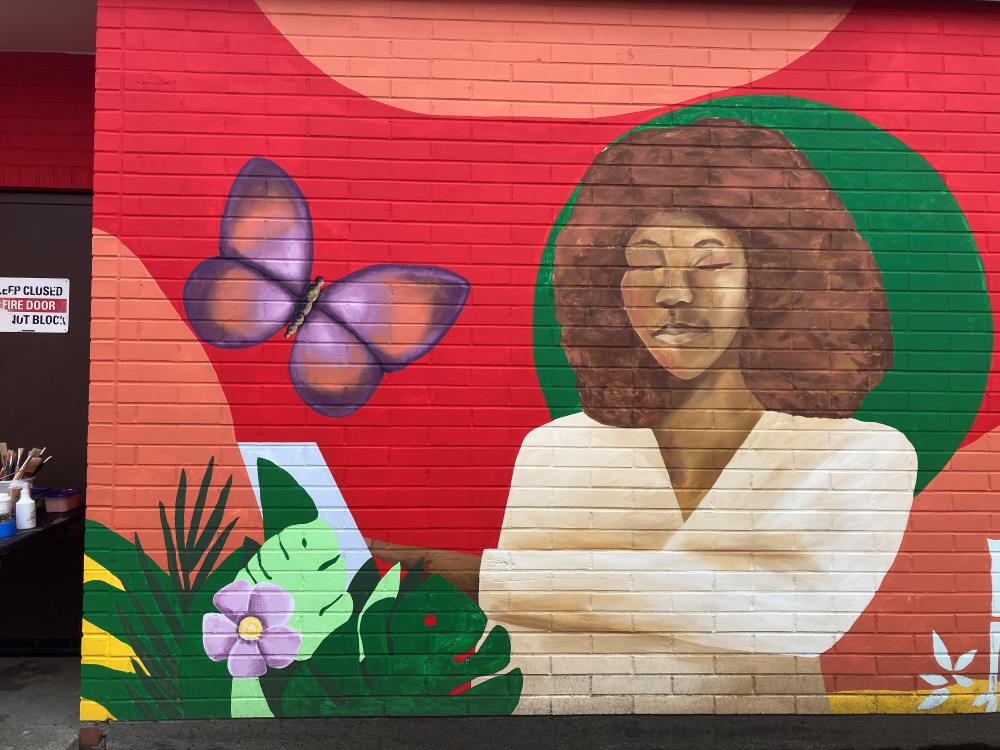 A woman pointing to a computer surrounded by bright colors of orange, red and green, tropical foliage and a purple and orange butterfly